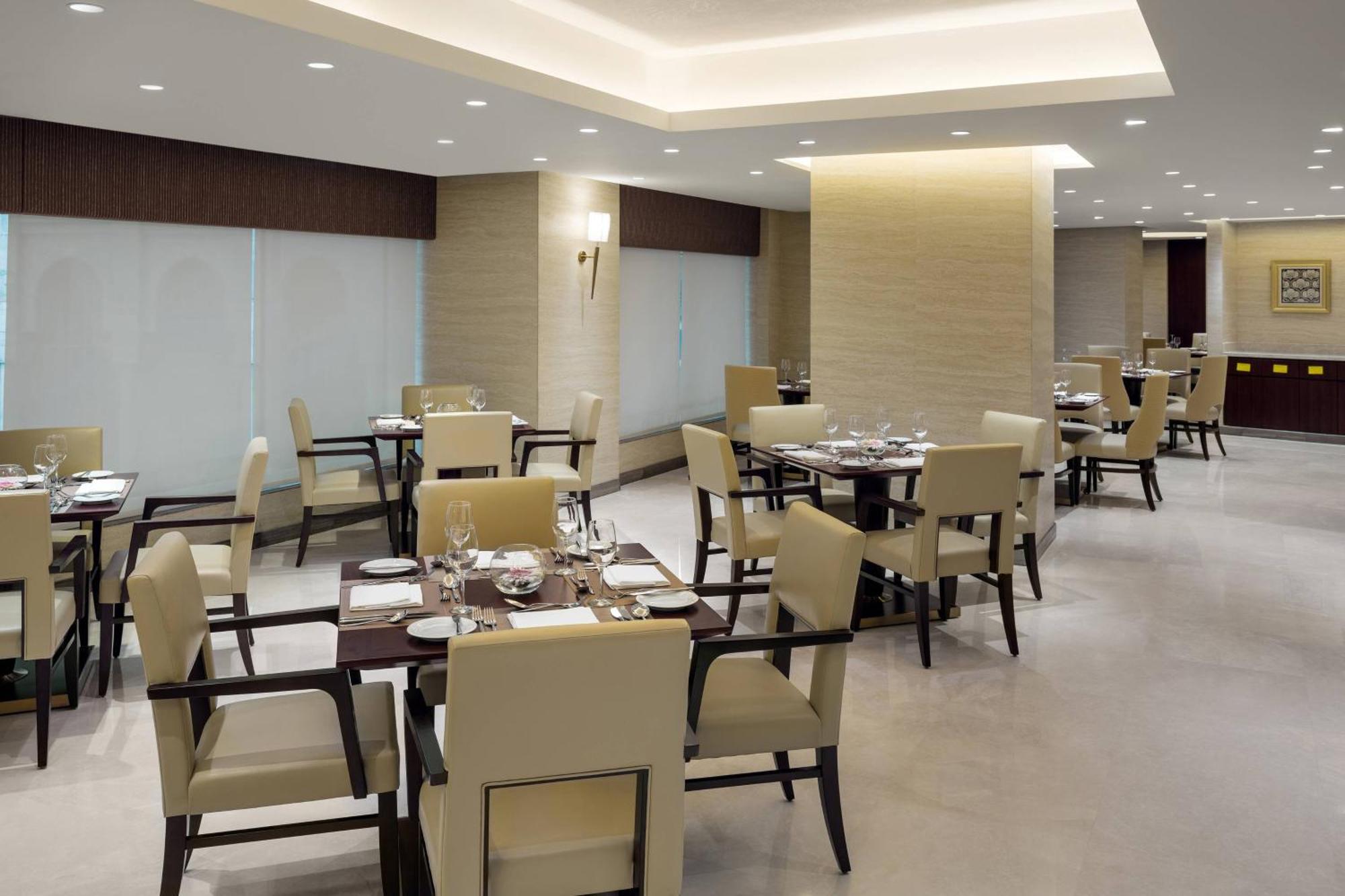 Sheraton Makkah Jabal Al Kaaba Hotel Мека Екстериор снимка The photo shows a modern restaurant interior. It features several dining tables, each set with utensils and tableware. The chairs are upholstered in a light fabric, and the overall decor is sleek and minimalistic, with soft lighting from overhead fix